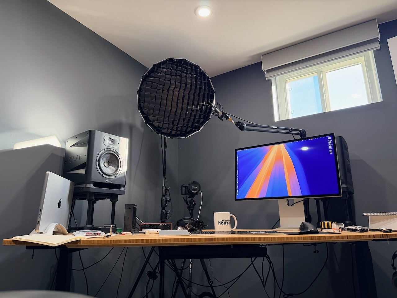 An image of a desk with a big speaker on it, as well as a computer setup. There's a camera pointing at the desk and a large studio light pointing where a person would naturally sit to work.